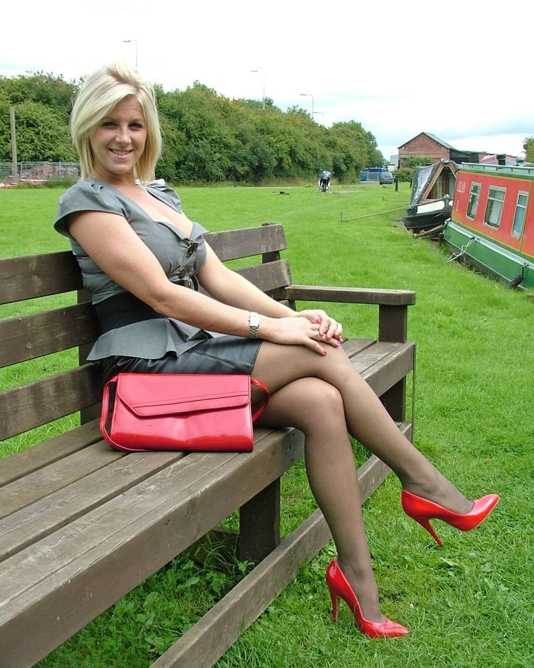 Clothed female with blonde hair shows off her red pumps in nylons on a bench
