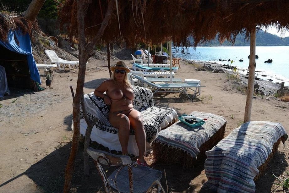 Thick blonde woman with big boobs wanders around a nude beach in a straw hat