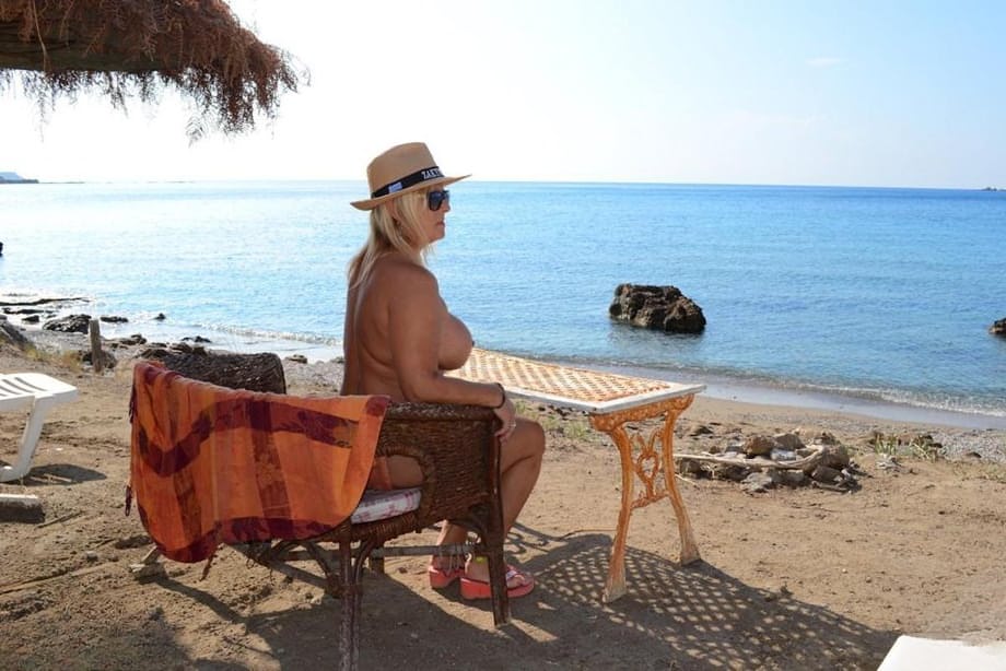 Thick blonde woman with big boobs wanders around a nude beach in a straw hat
