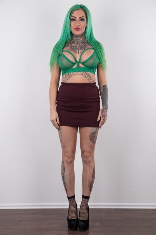 Tattooed girl with green hair and pierced nipples stands naked after disrobing