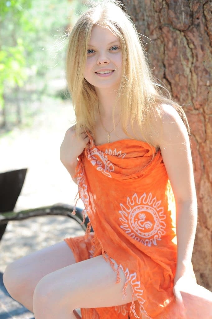 Young blonde Kisa removes her wrap to pose nude on bench under an old tree