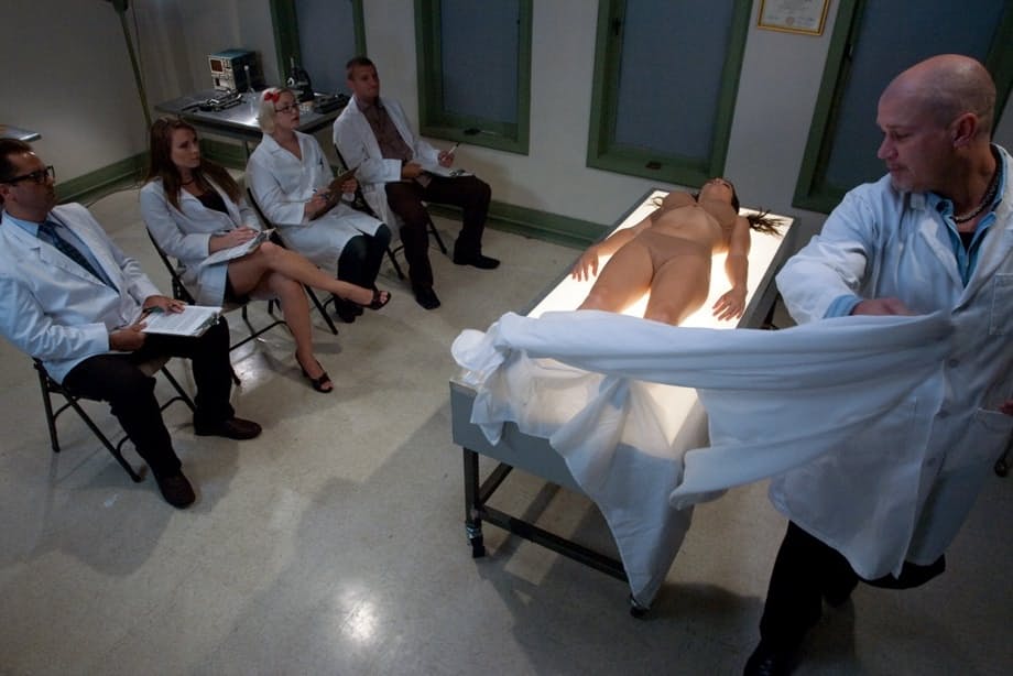 Bald doctor examines naked body of good-looking assistant in people's presence